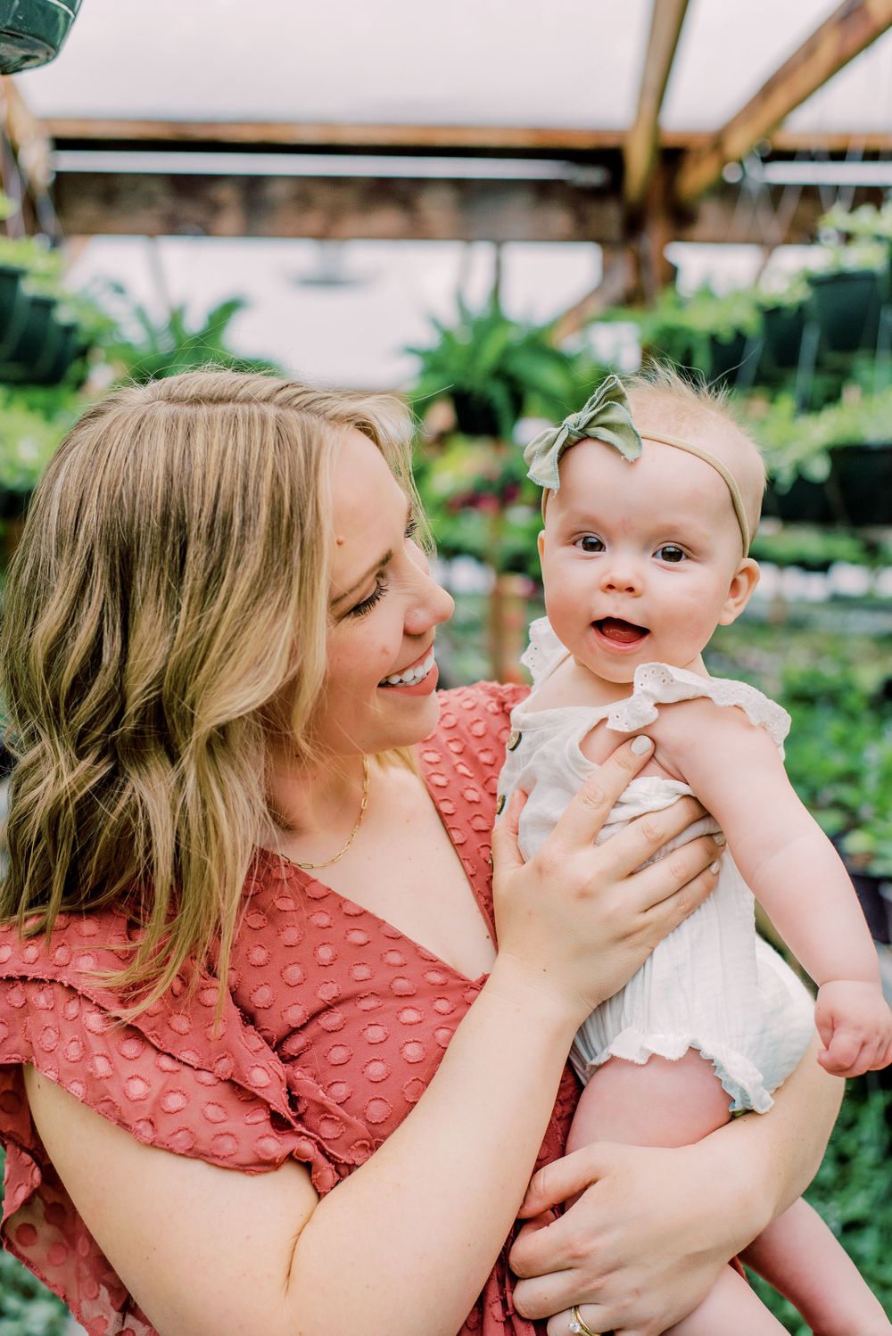 A tender moment captured between a woman in a coral dress and baby in white outfit among lush greenery in a garden setting.