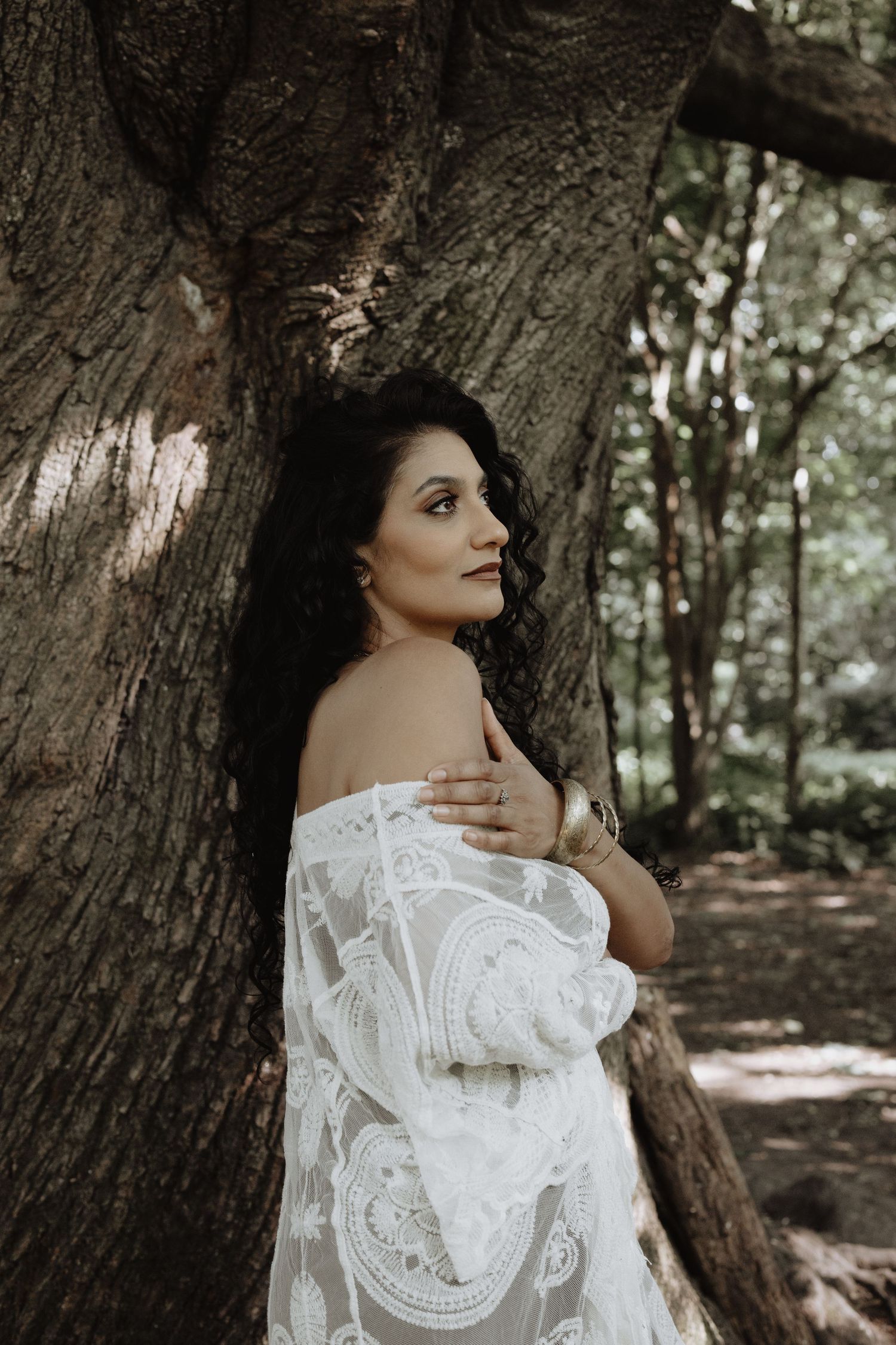 Artistic portrait of a woman in white lace against a large tree trunk in a forest setting.
