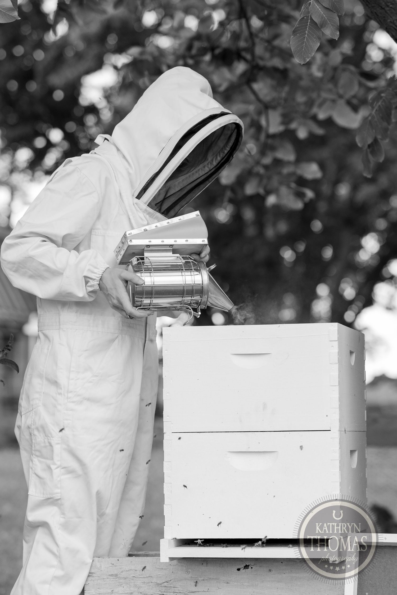 Beekeeper in protective gear working with equipment at a white beehive box in black and white.