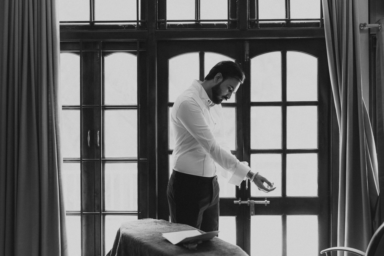 A figure in white adjusts clothing while standing by tall windows with curtains.