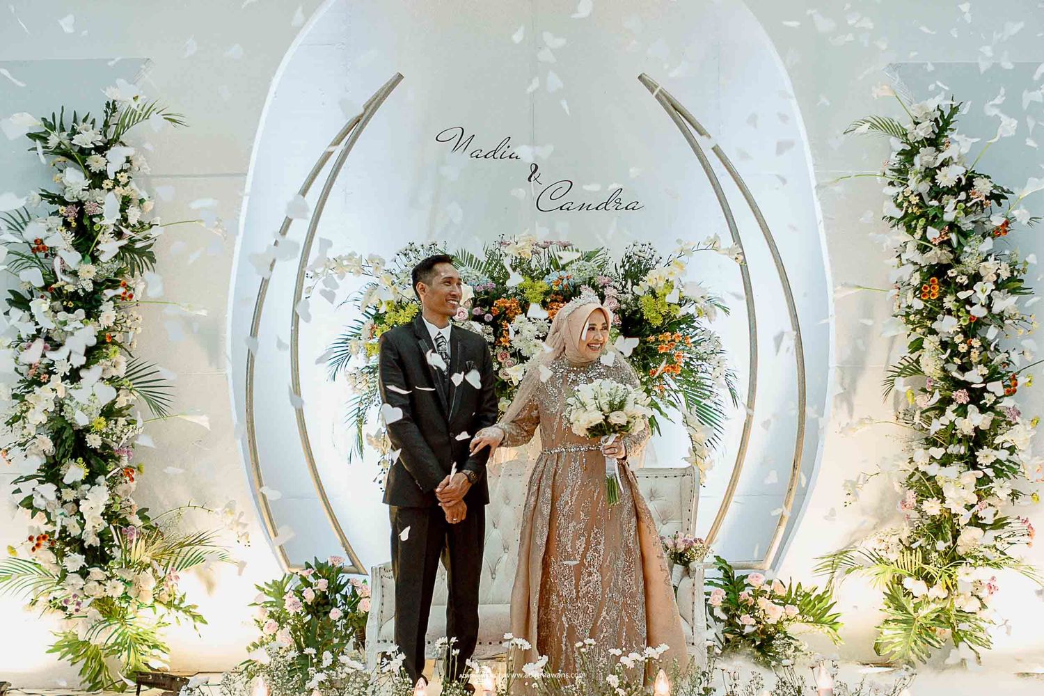 Newlyweds pose under elegant white circular arch decorated with fresh flowers.