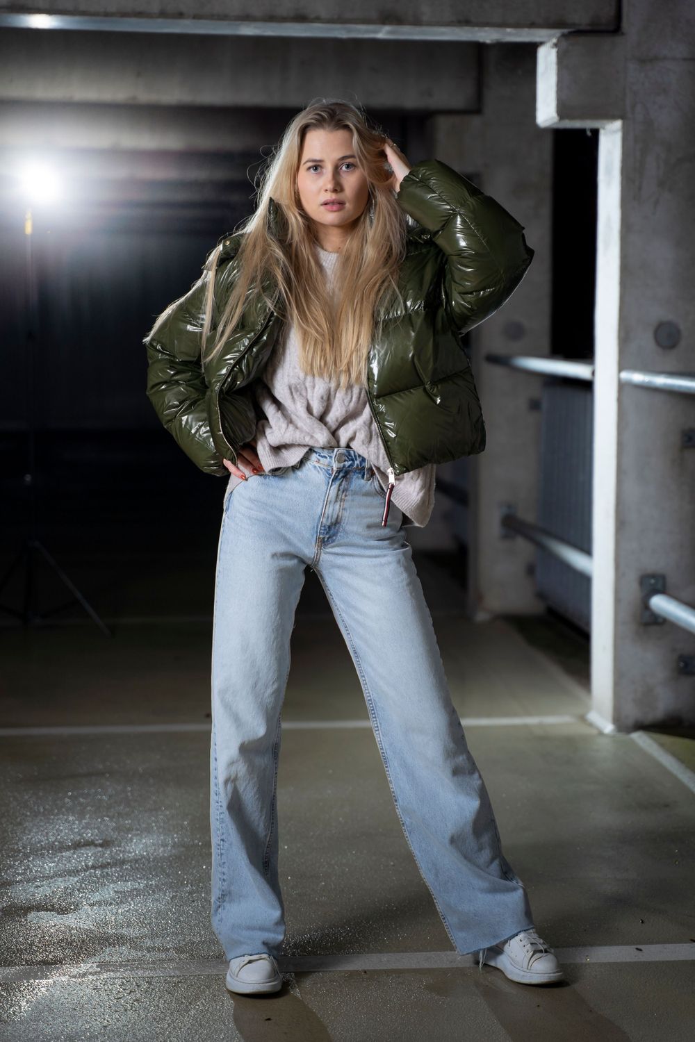 Fashion model wearing an olive green puffer jacket and light wash jeans in an industrial setting.