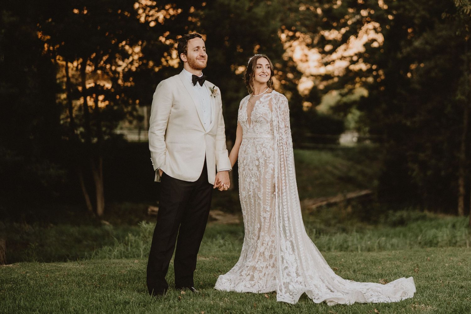 A couple stands together in formal wedding attire on a grassy lawn at sunset.