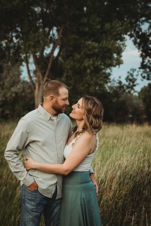 A romantic couple embraces in a sunlit meadow with tall grass and trees in the background during golden hour.