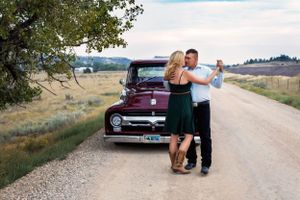 Couple dances next to a vintage red pickup truck on a rural dirt road surrounded by countryside.