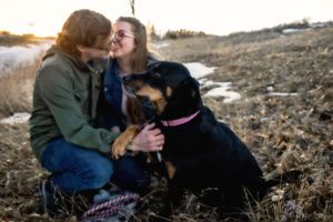 A couple sits outdoors on a hillside embracing their black dog during golden hour.