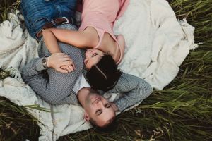 Two people lying together on a white blanket in tall grass, sharing a romantic moment while looking upward.