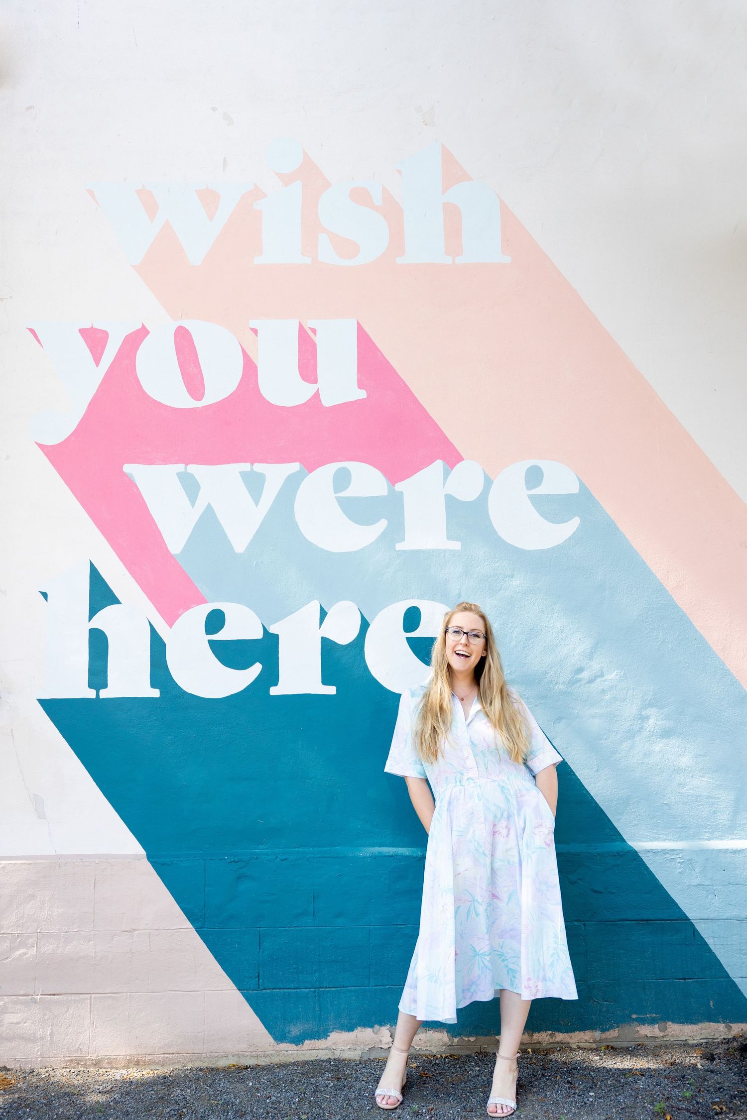 A person in a white dress poses against a colorful wall mural that reads 'wish you were here'.