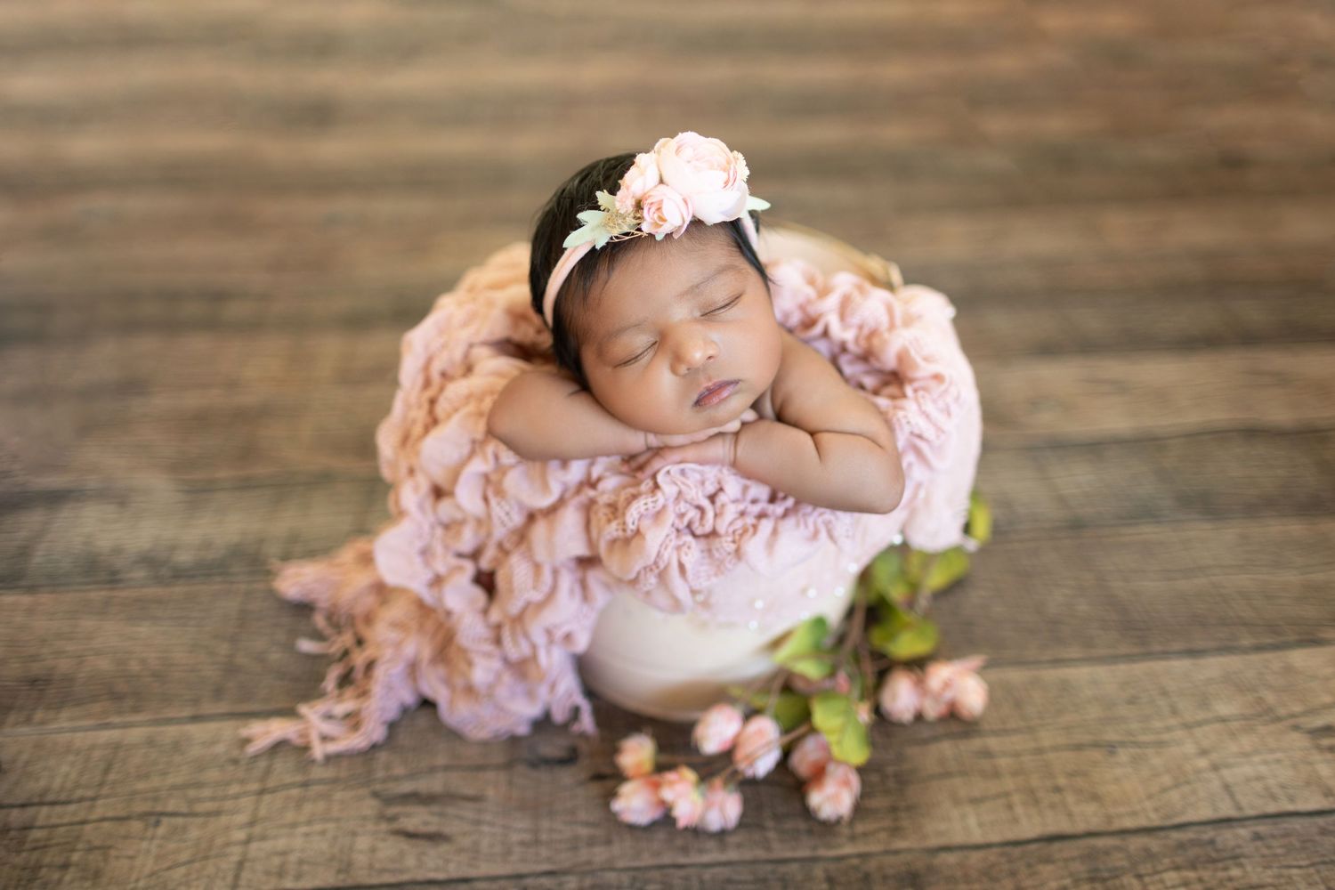 Sleeping newborn in pink lace wrap with floral crown on wooden backdrop.