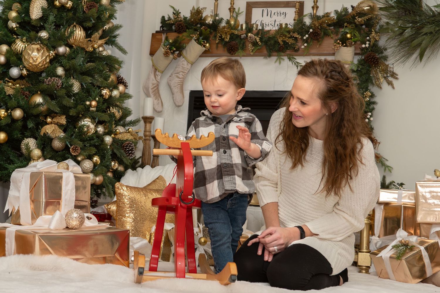 Parent and toddler play with red rocking horse near Christmas tree and wrapped presents.