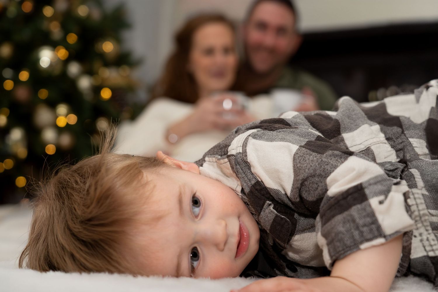 Close up of toddler in plaid shirt lying down with Christmas tree in background.