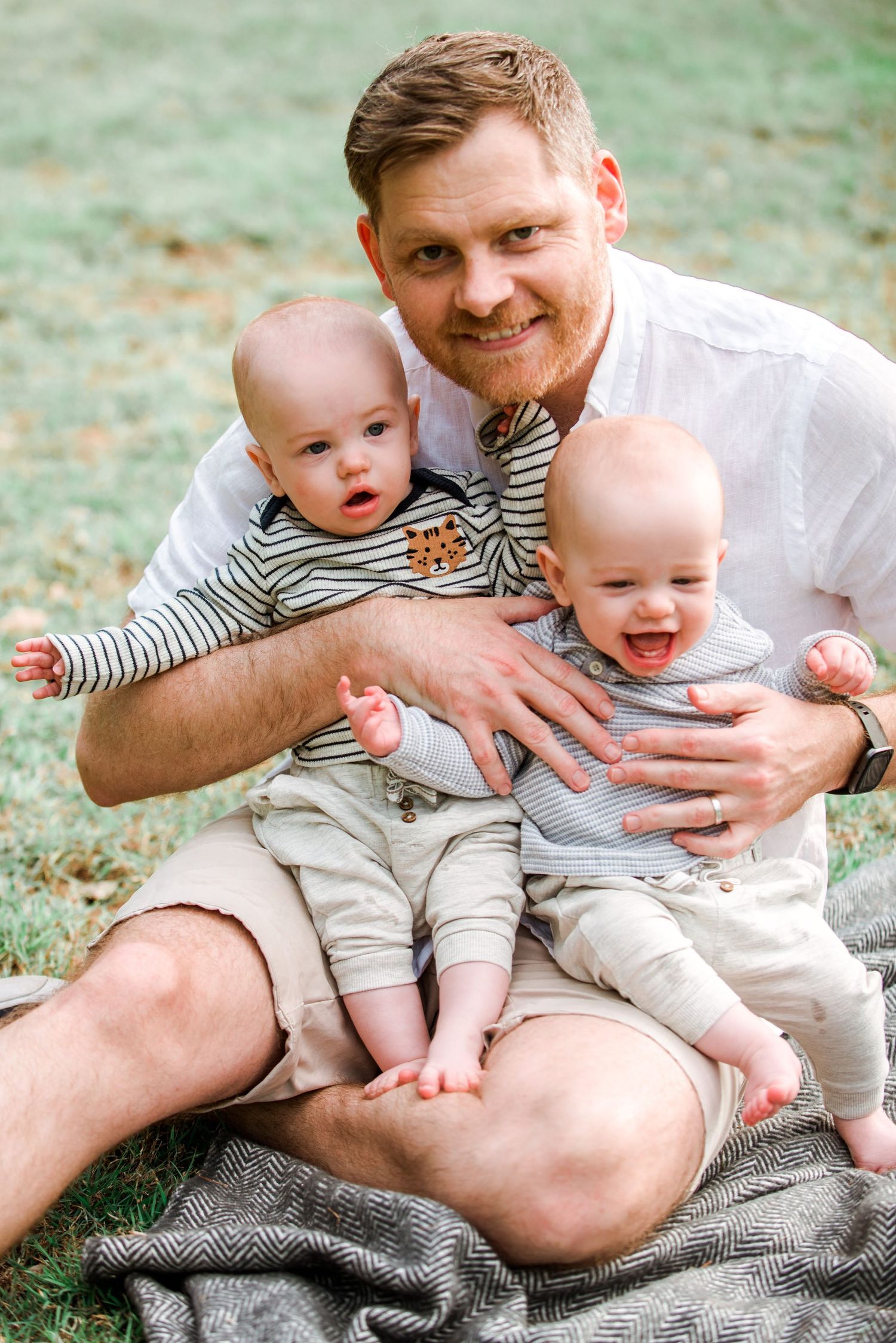 Two adorable infants in striped outfits sit with an adult on a grey blanket in an outdoor setting.