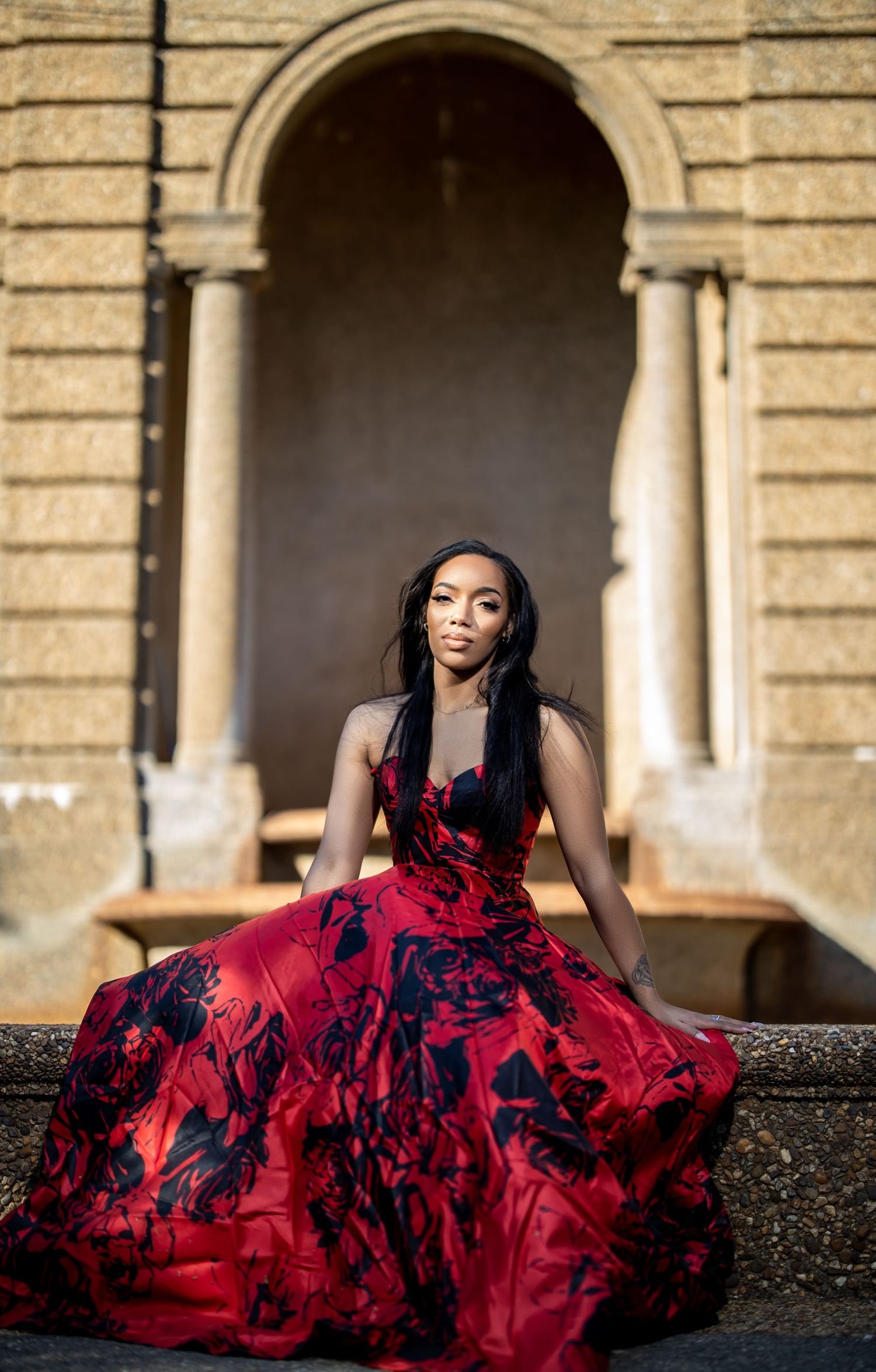 A person in a red and black floral ball gown poses in front of an ornate stone archway.
