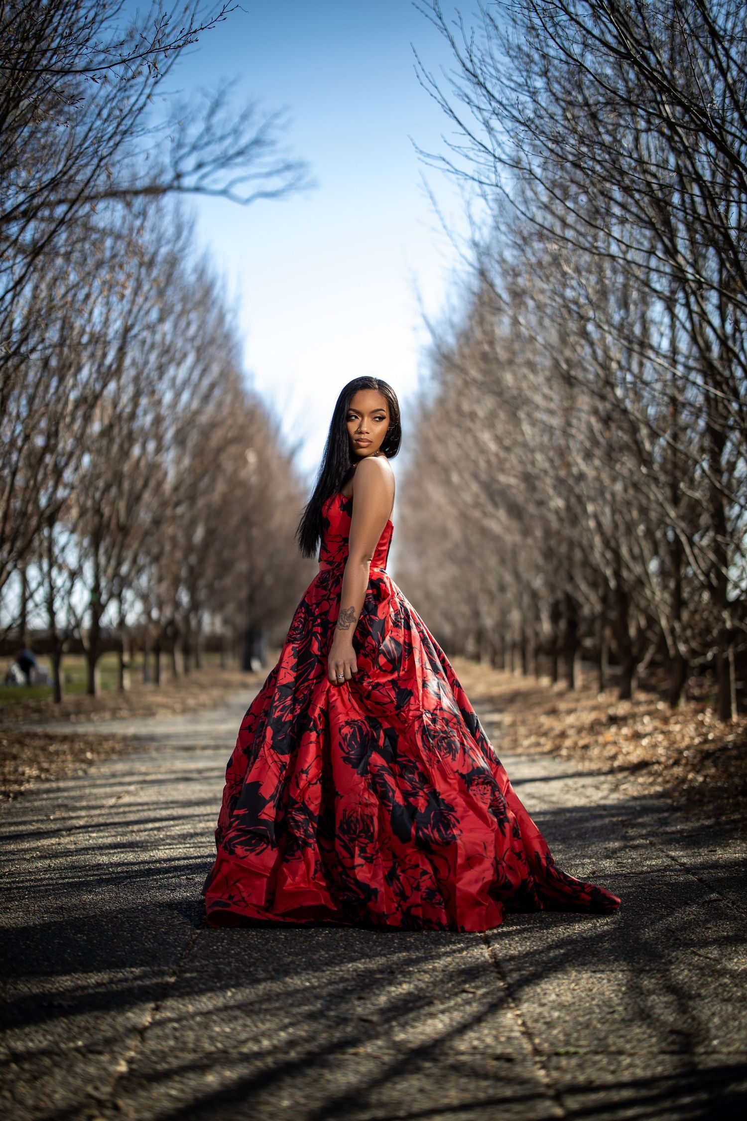 A person in a red floral gown poses on a tree-lined path during winter.