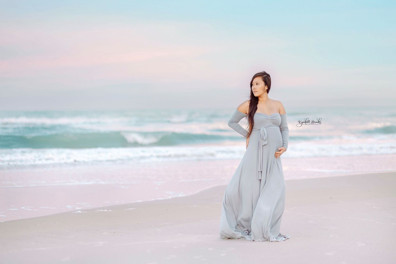 Woman in flowing grey dress poses elegantly on a pastel beach at sunset.