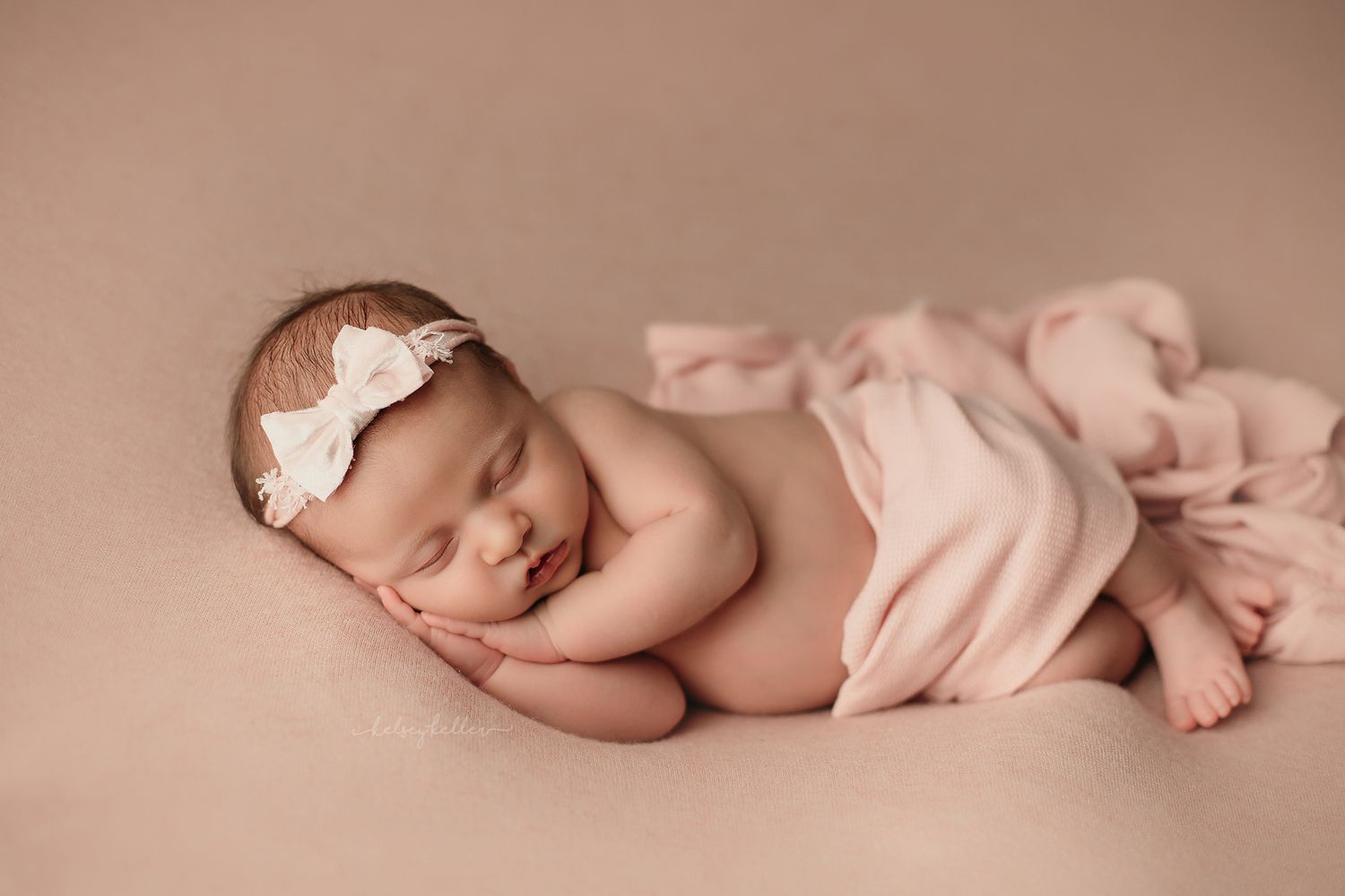 A sleeping newborn baby wearing a delicate headband lies peacefully on soft pink fabric.