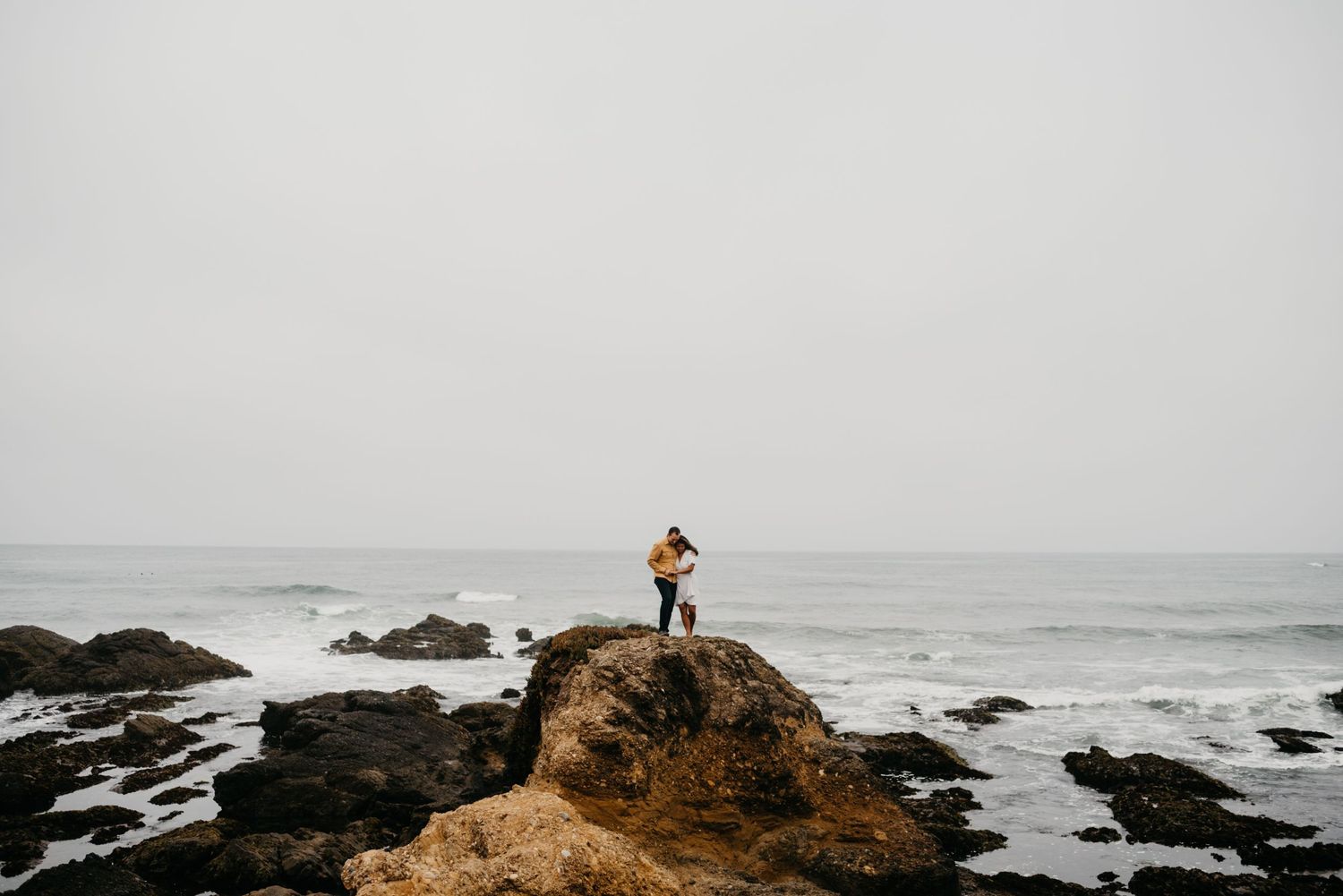 Engaged couple embracing on rocky cliff overlooking the Pacific ocean