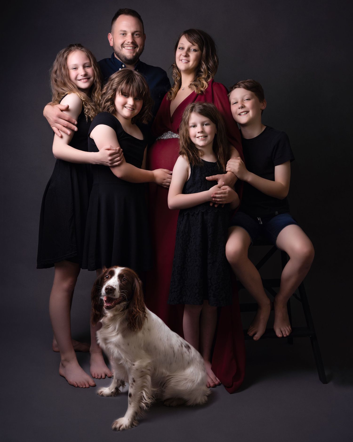 Family portrait with parents, children and their smiling spaniel dog against dark backdrop.