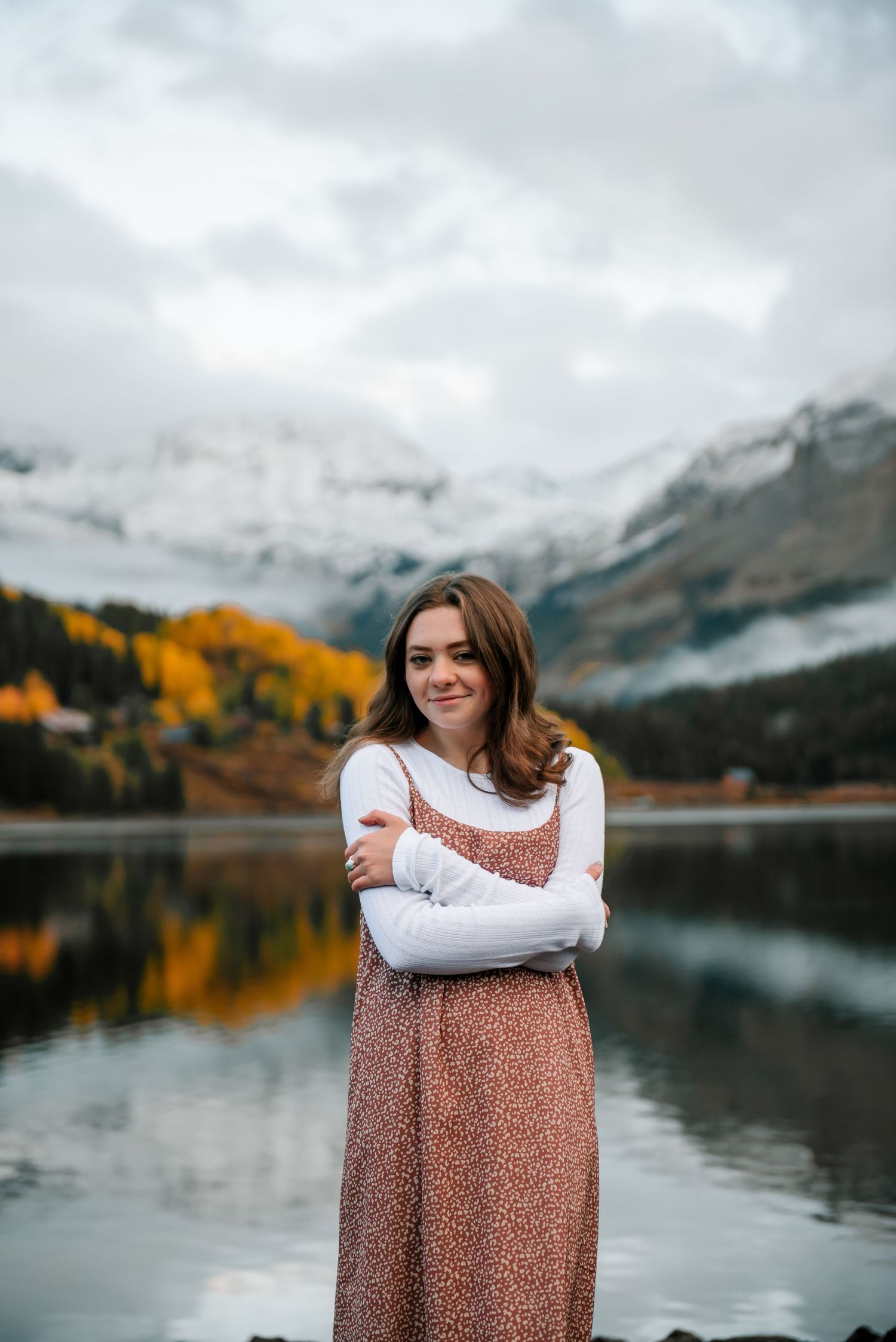 Person in long dress hugs themselves while standing by scenic mountain lake with autumn colors.