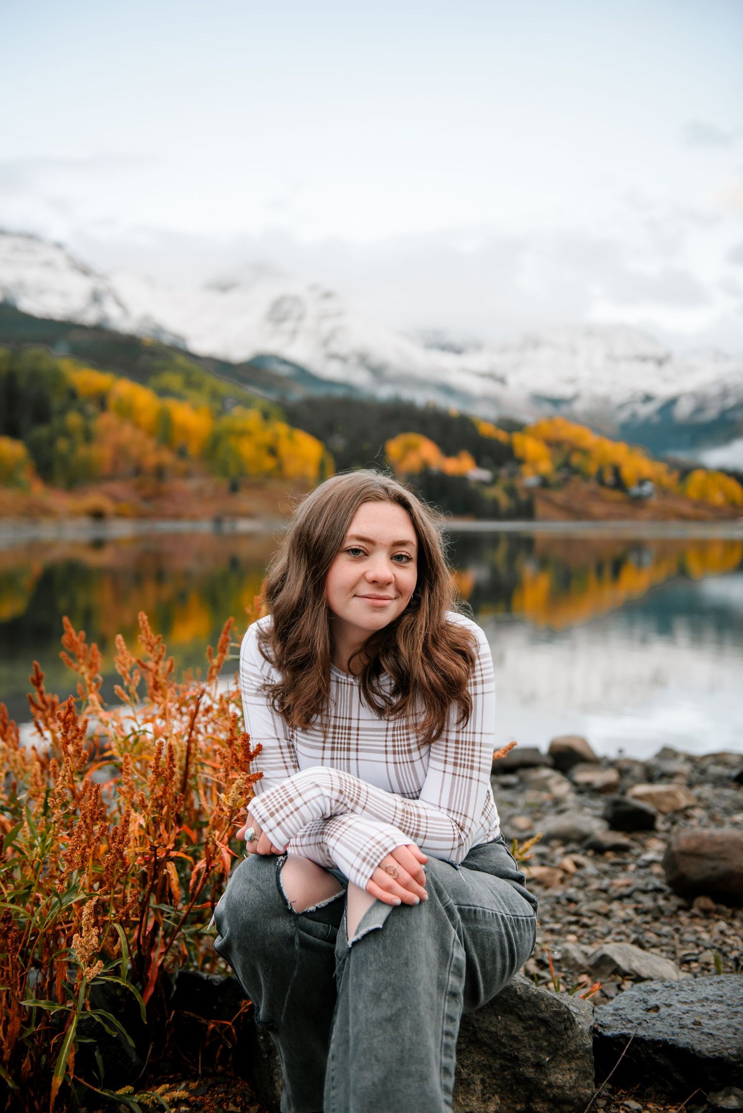 Person in plaid shirt sits on rocky shoreline with fall colors and snowy mountains in background.