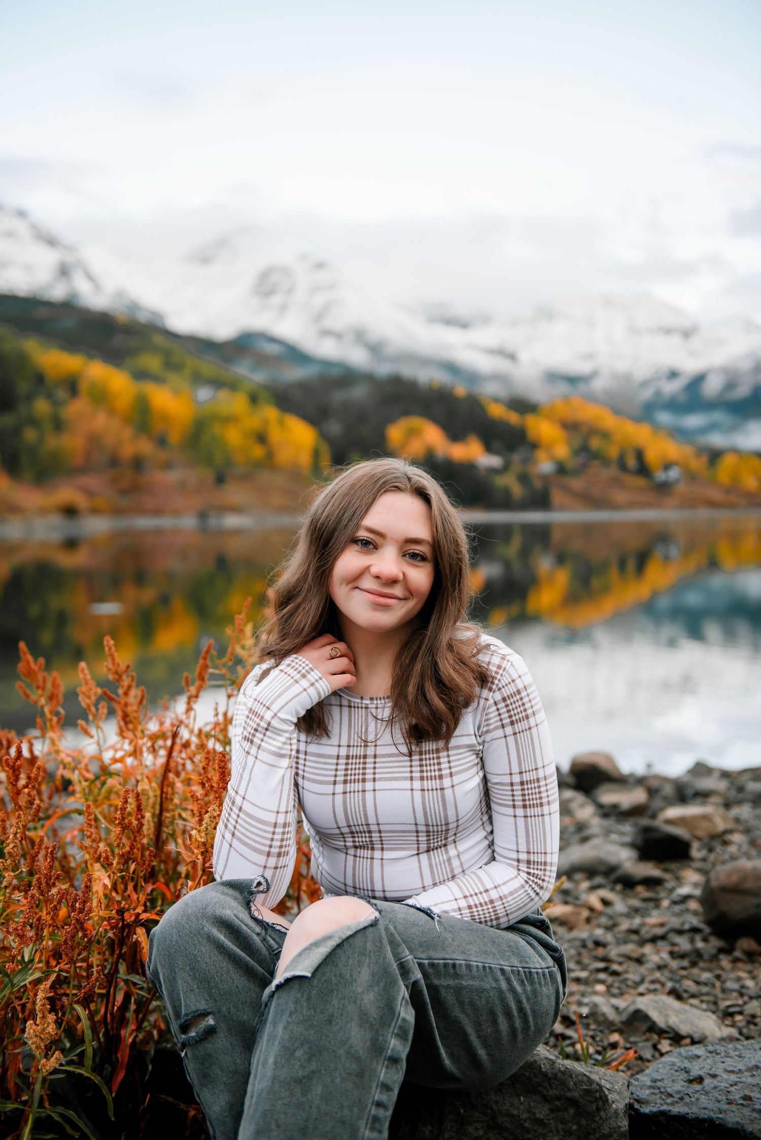 Person in casual attire sits by lakeside with orange fall foliage and mountain reflections in water.