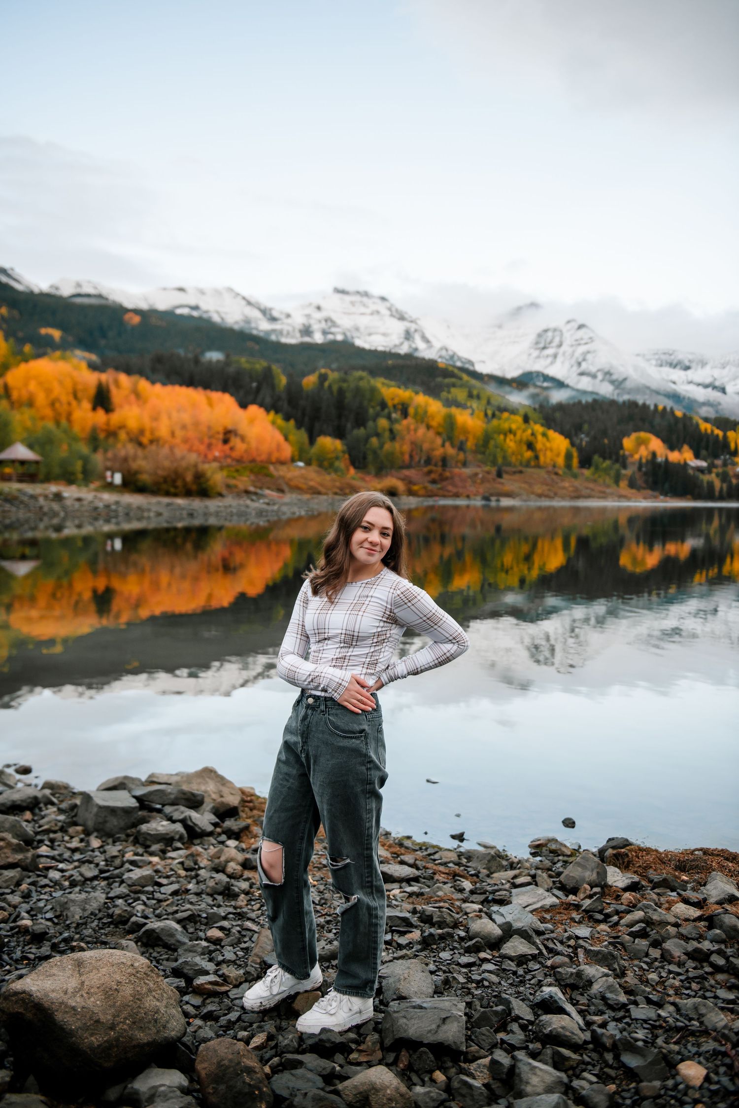 Person poses on rocky lakeshore with snow-capped mountains and golden autumn trees reflecting in water.