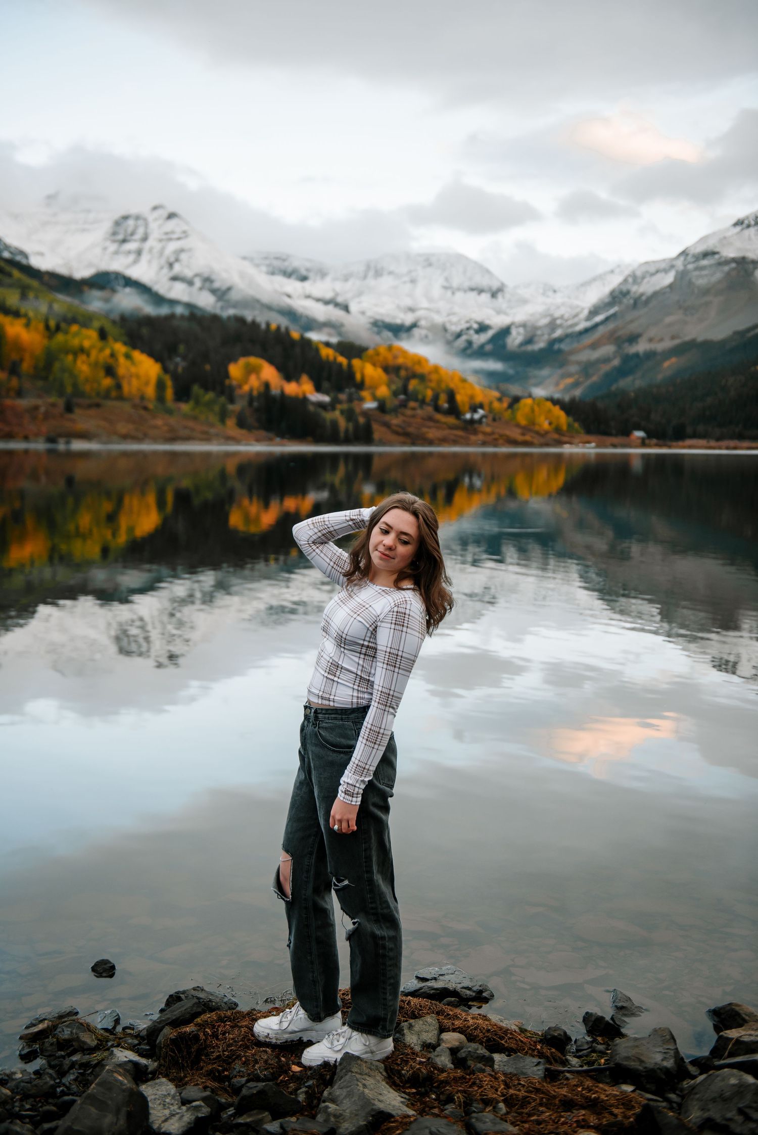 Person in plaid top poses playfully by mountain lake with dramatic fall scenery reflections.