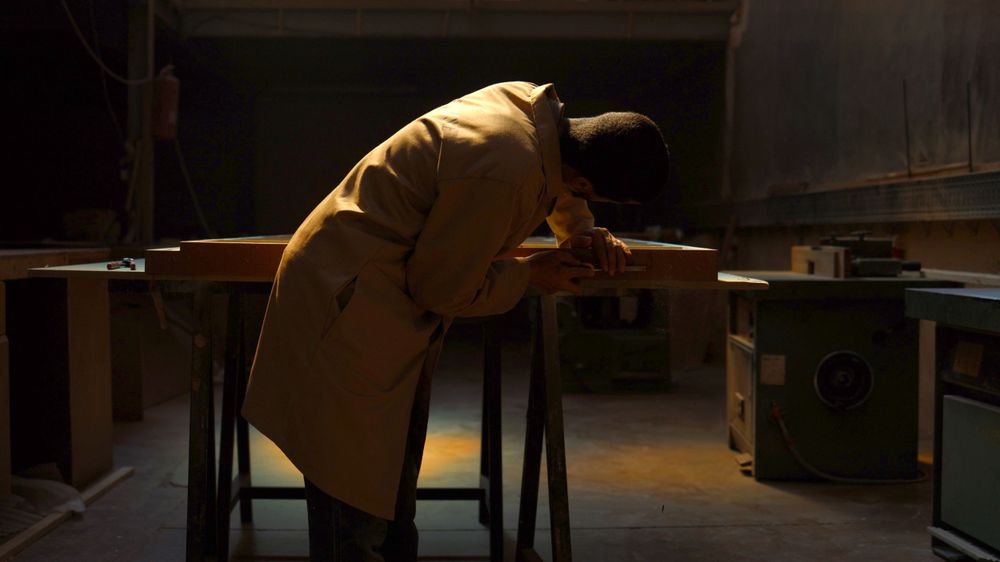 A figure in a beige coat leans over a desk in a dimly lit room.