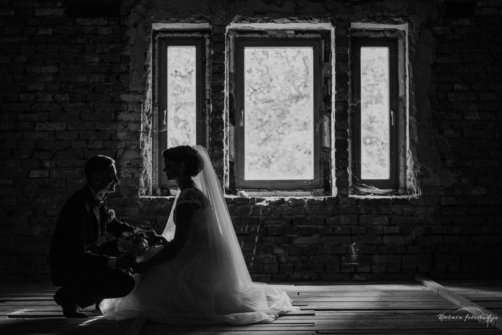 juodai balta vestuvinė fotosesija, jaunieji susikabinę sėdi tamsoje priešais langą , žiūrėdami vienas į kitą