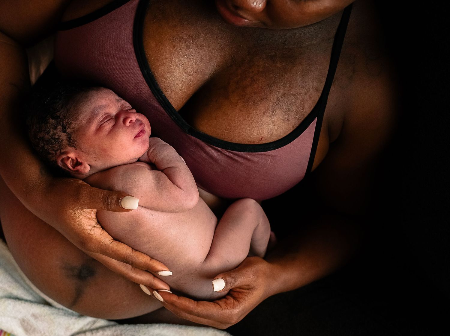 black woman in purple top holding sleeping newborn on bare pregnant belly
