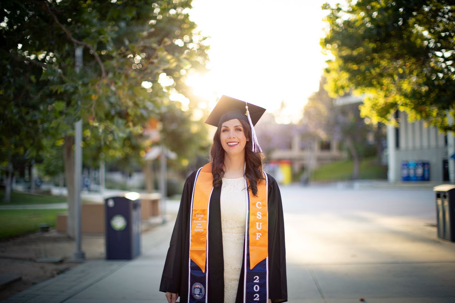 Graduate in cap and gown smiling during golden hour on campus walkway.