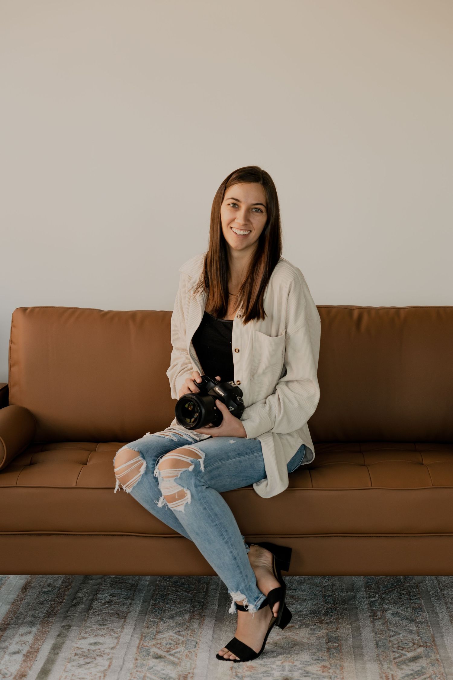 Photographer relaxes on brown leather couch in distressed jeans and white cardigan with camera.