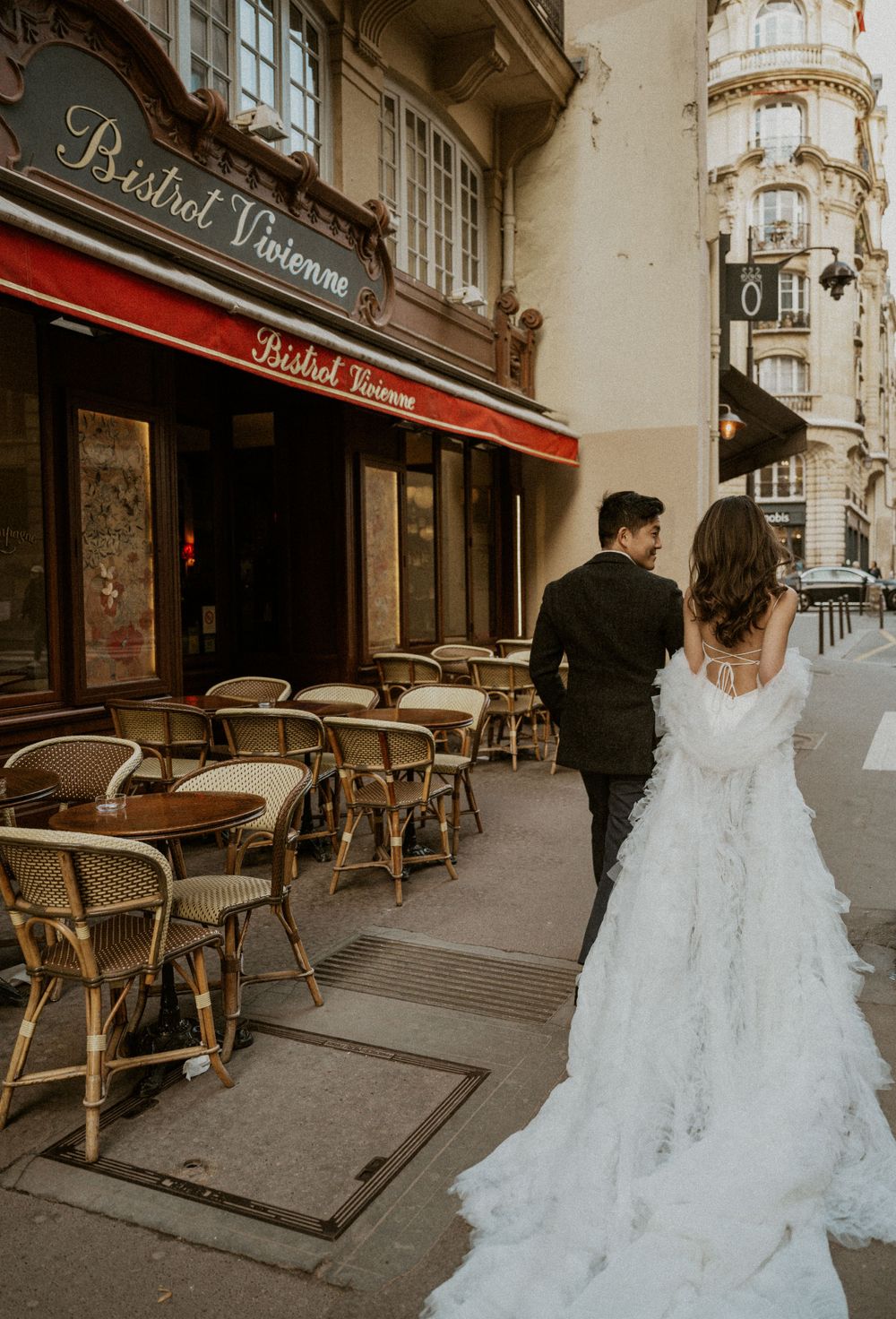 A couple in wedding attire stands near a Parisian bistro with classic rattan chairs and red awning.