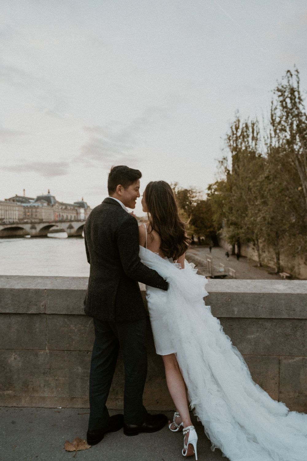 Couple in formal attire embraces against Parisian riverside backdrop at sunset.