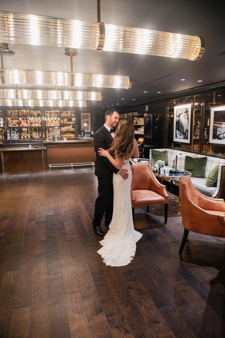 An elegant wedding couple embraces in a luxurious bar setting with warm lighting and leather chairs.