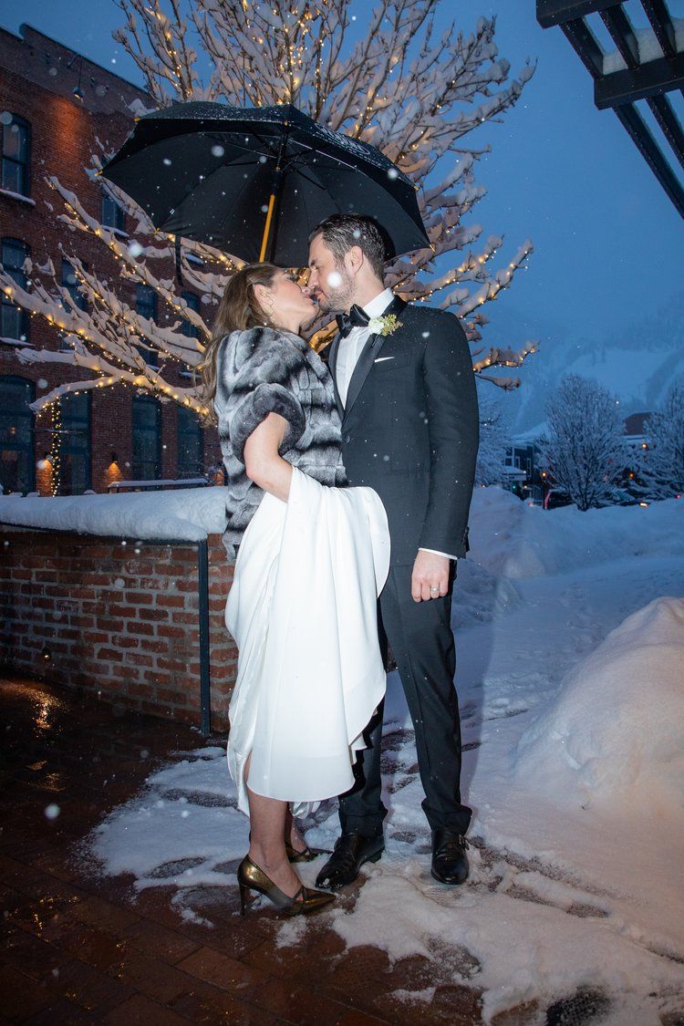 A romantic winter wedding moment under an umbrella with falling snow.