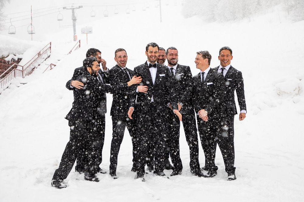 A group in black suits pose together in falling snow during a winter wedding celebration.