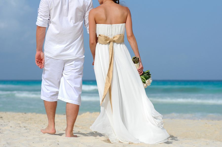 Couple in white attire standing on a beach holding hands with ocean waves in background.