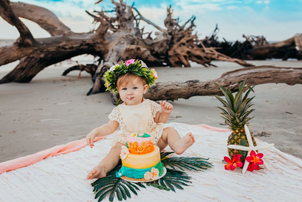 Adorable baby girl in a floral crown celebrates first birthday with a tropical cake on the beach.
