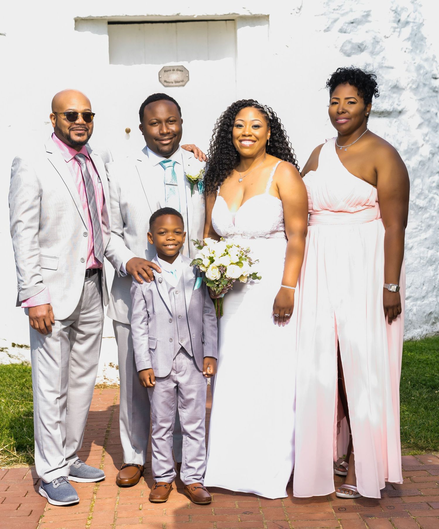A wedding party in coordinating light-colored attire poses together against a white building.