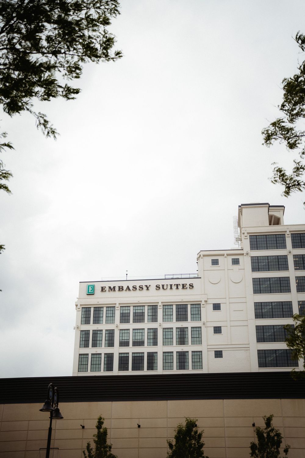 Embassy Suites hotel exterior with white facade and trees framing the building on a cloudy day.