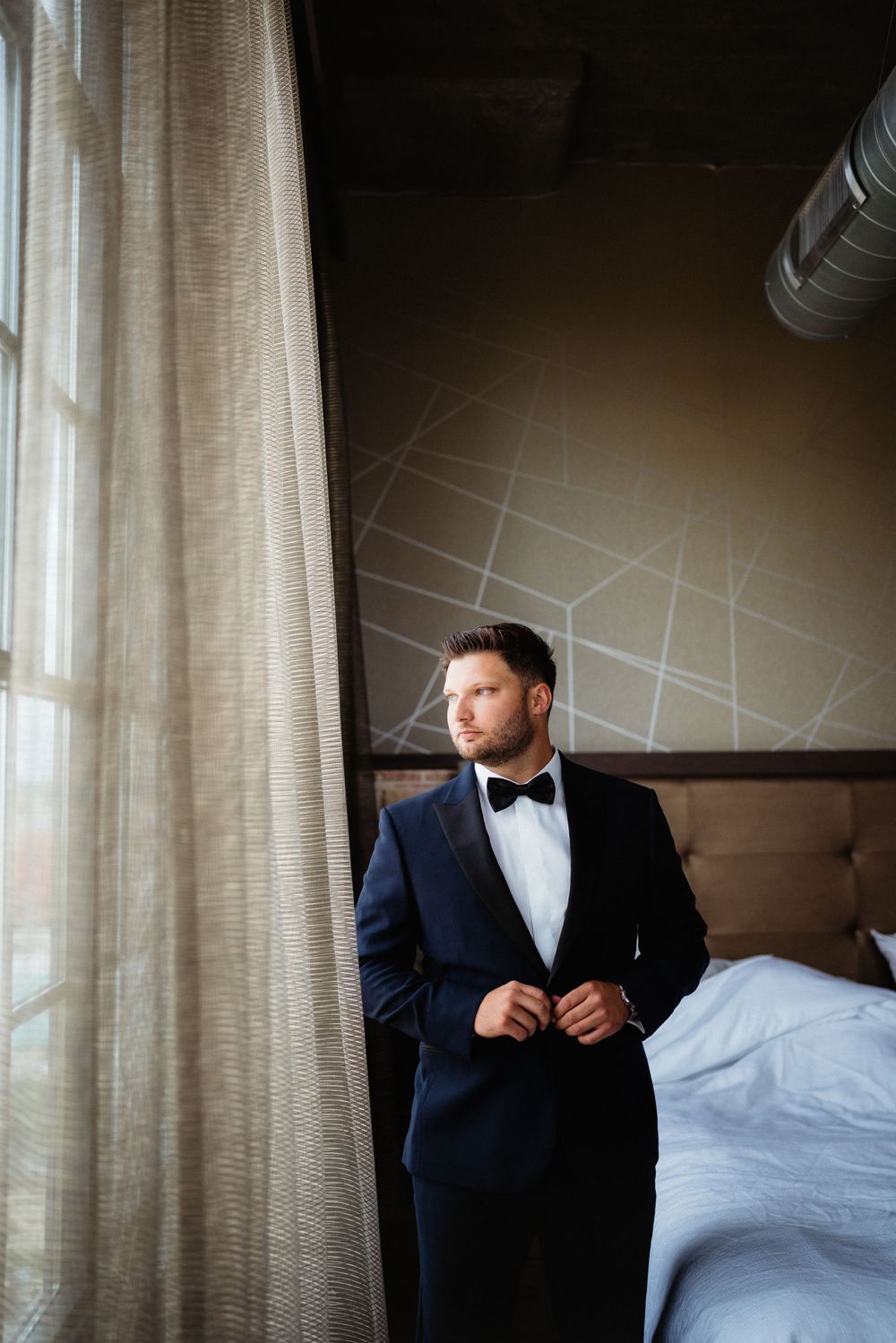 Elegant groom in navy tuxedo and bow tie stands by window in hotel room.
