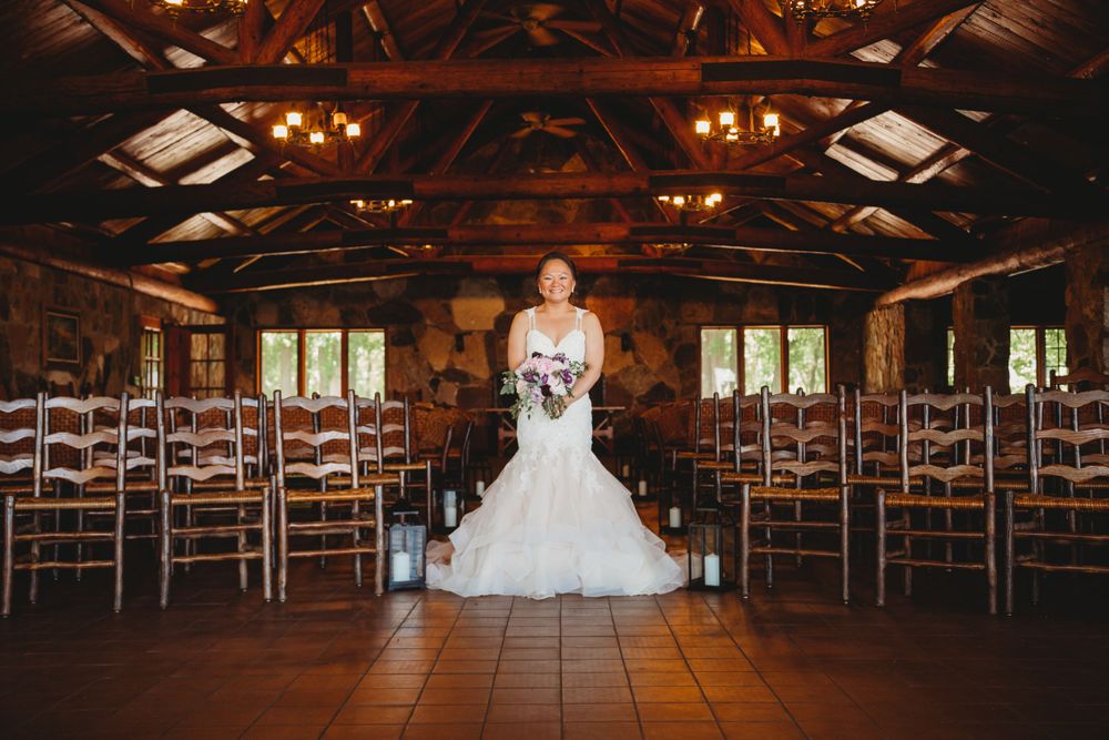 A bride in a white dress stands alone in a rustic wooden chapel with rows of empty chairs.