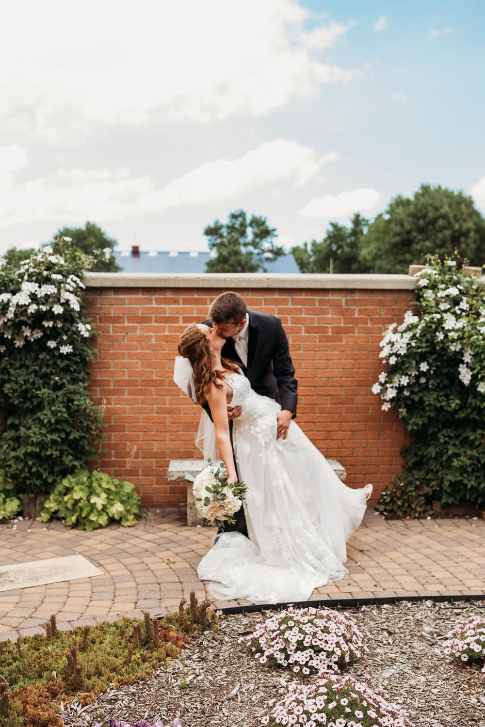 Romantic wedding couple posing against a brick wall surrounded by white flowering bushes.