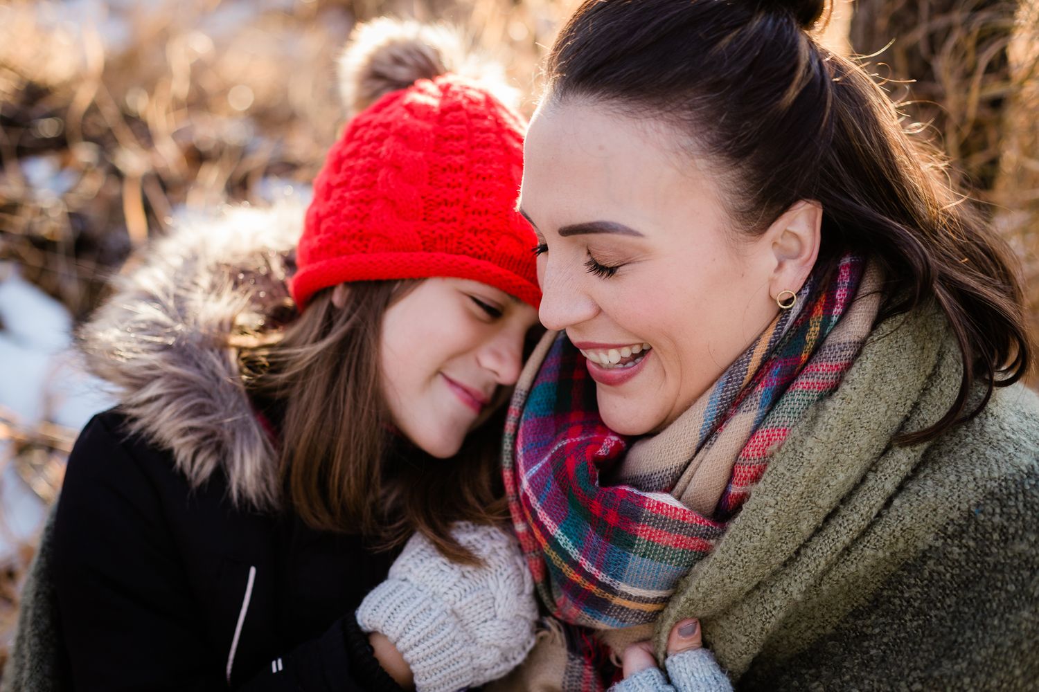 Two young women share a joyful winter embrace outdoors, one wearing a red knit hat and fur-trimmed coat.
