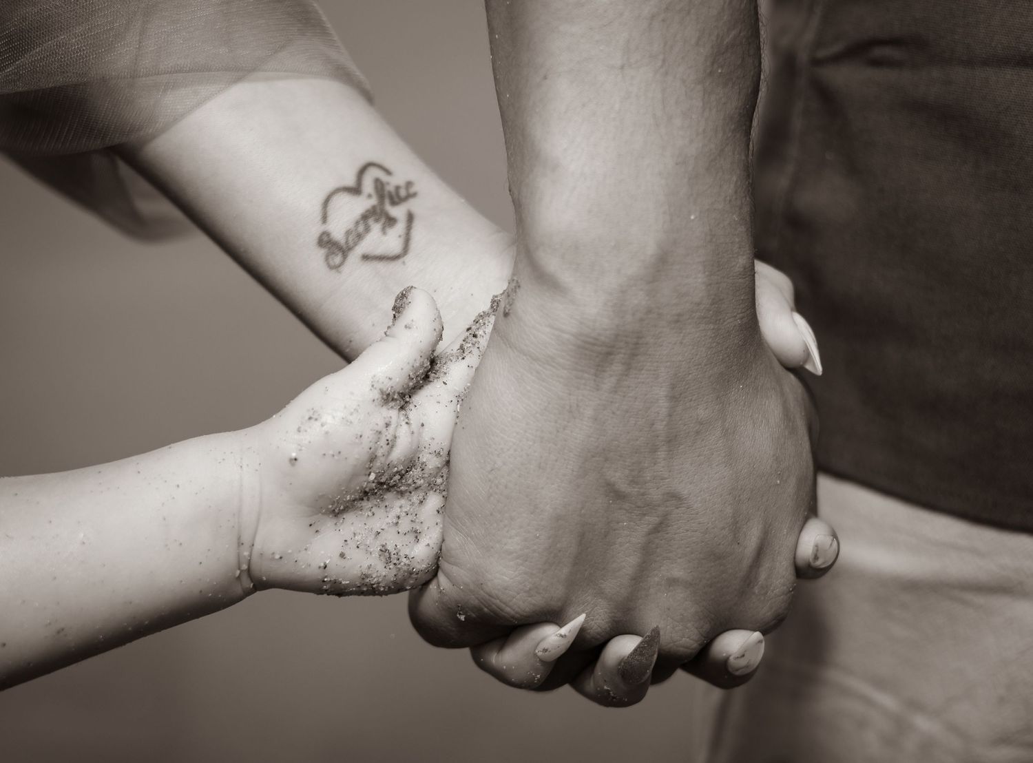A black and white close-up photograph of two people holding hands with a small anchor tattoo visible.