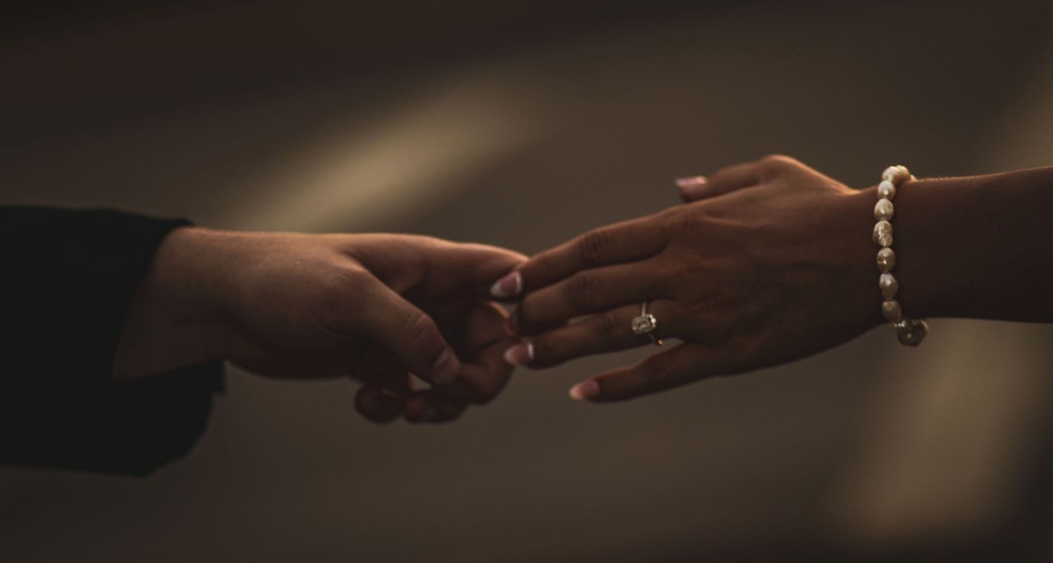 Close-up of hands exchanging engagement ring with bracelet detail in dim lighting.