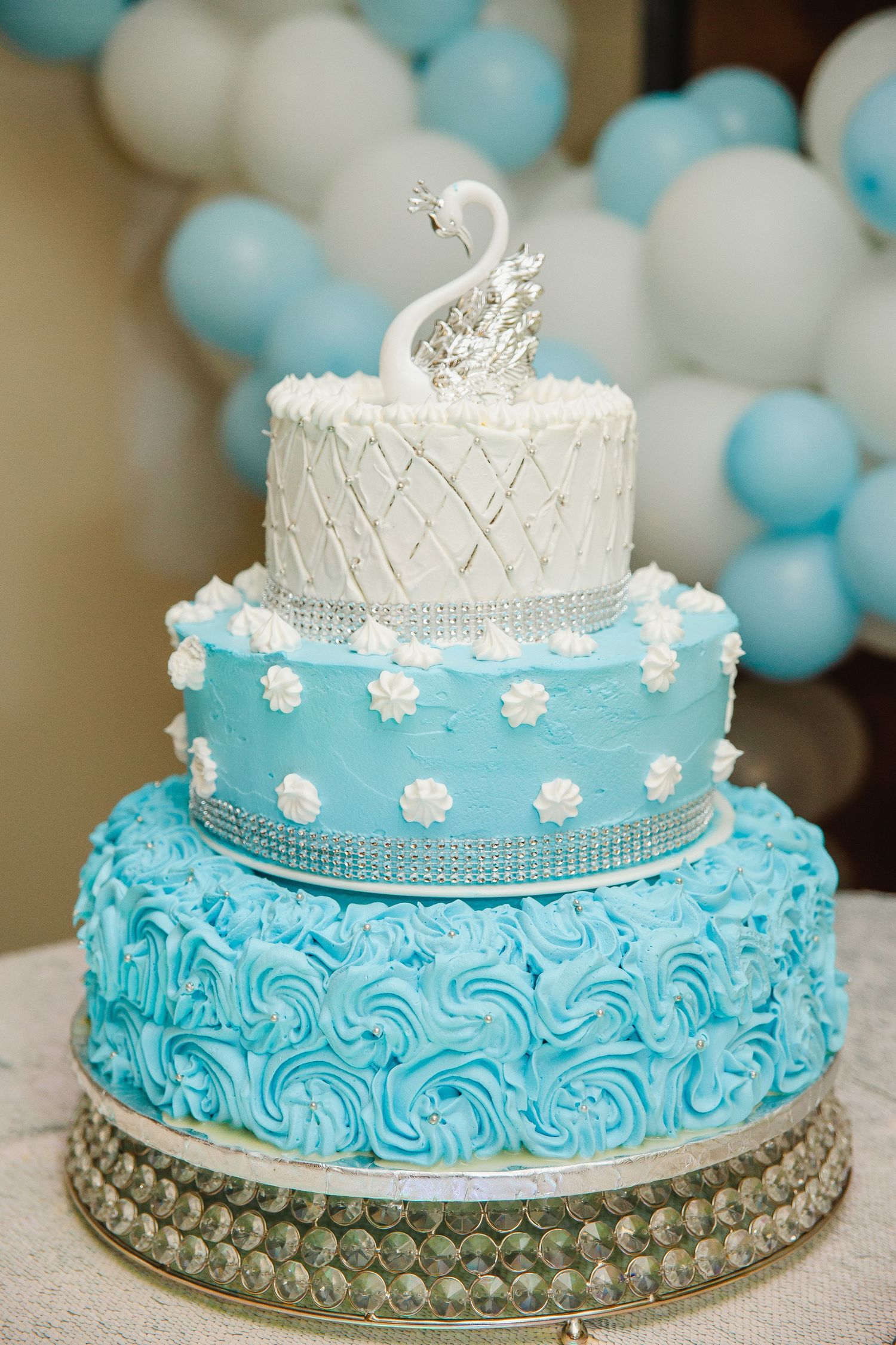 Three-tiered turquoise and white wedding cake with silver crown topper and rhinestone details.