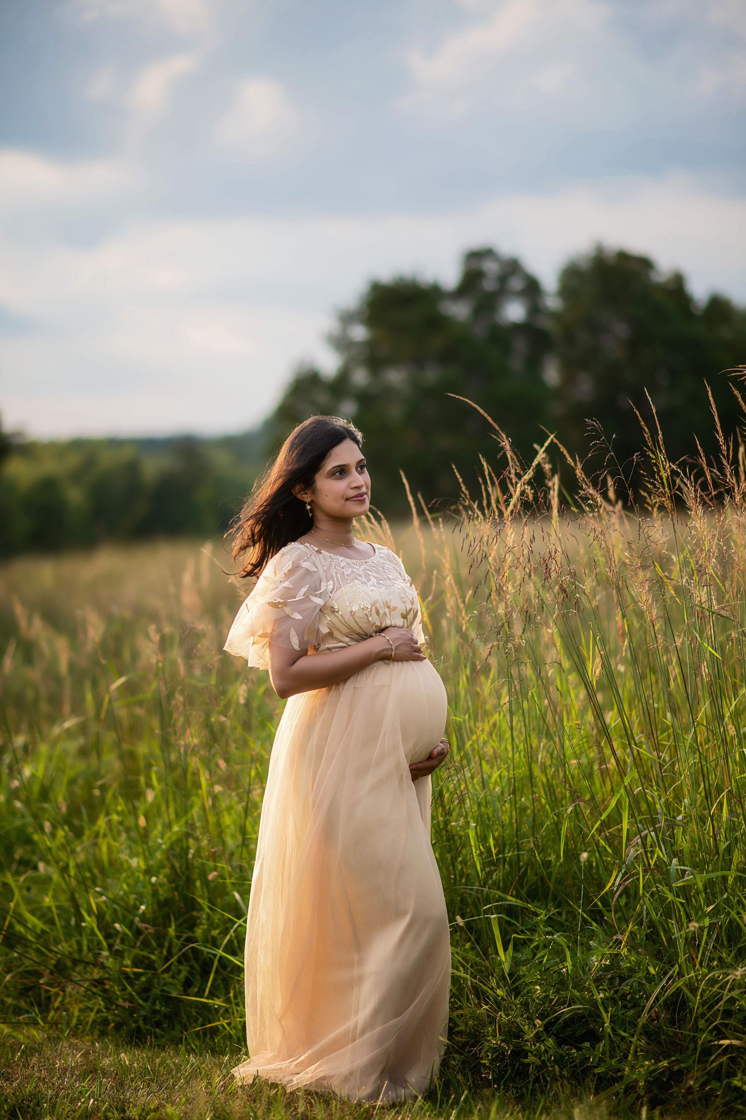 Woman in vintage white dress stands among tall prairie grass during golden hour.