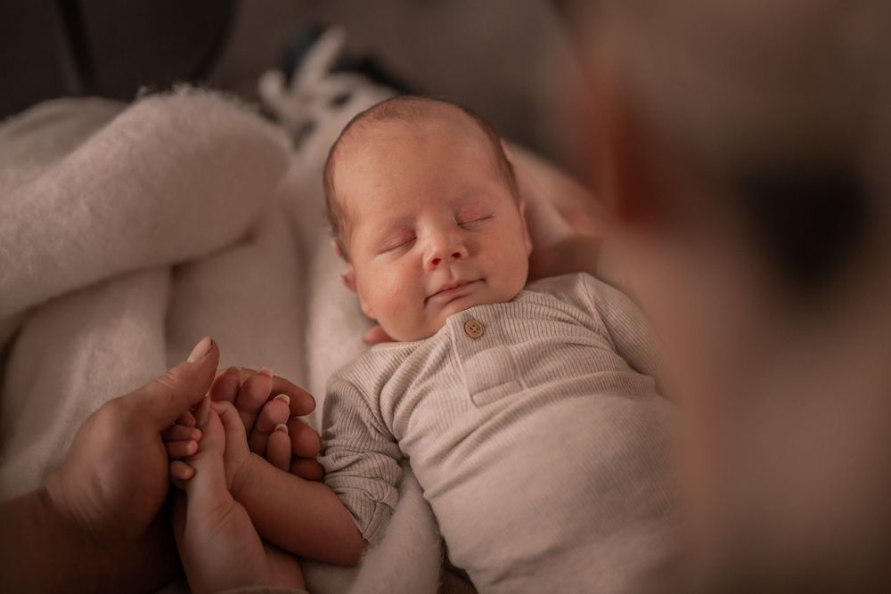 En fredeligt sovende nyfødt baby i hvid onesie holder hånd med en person i blød, varm belysning.